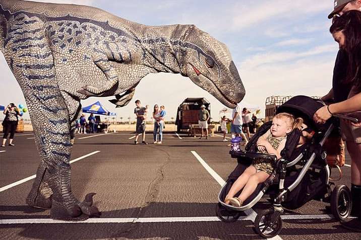 Child in a stroller gazes at a large dinosaur figure in an outdoor setting with other people in the background on a sunny day.