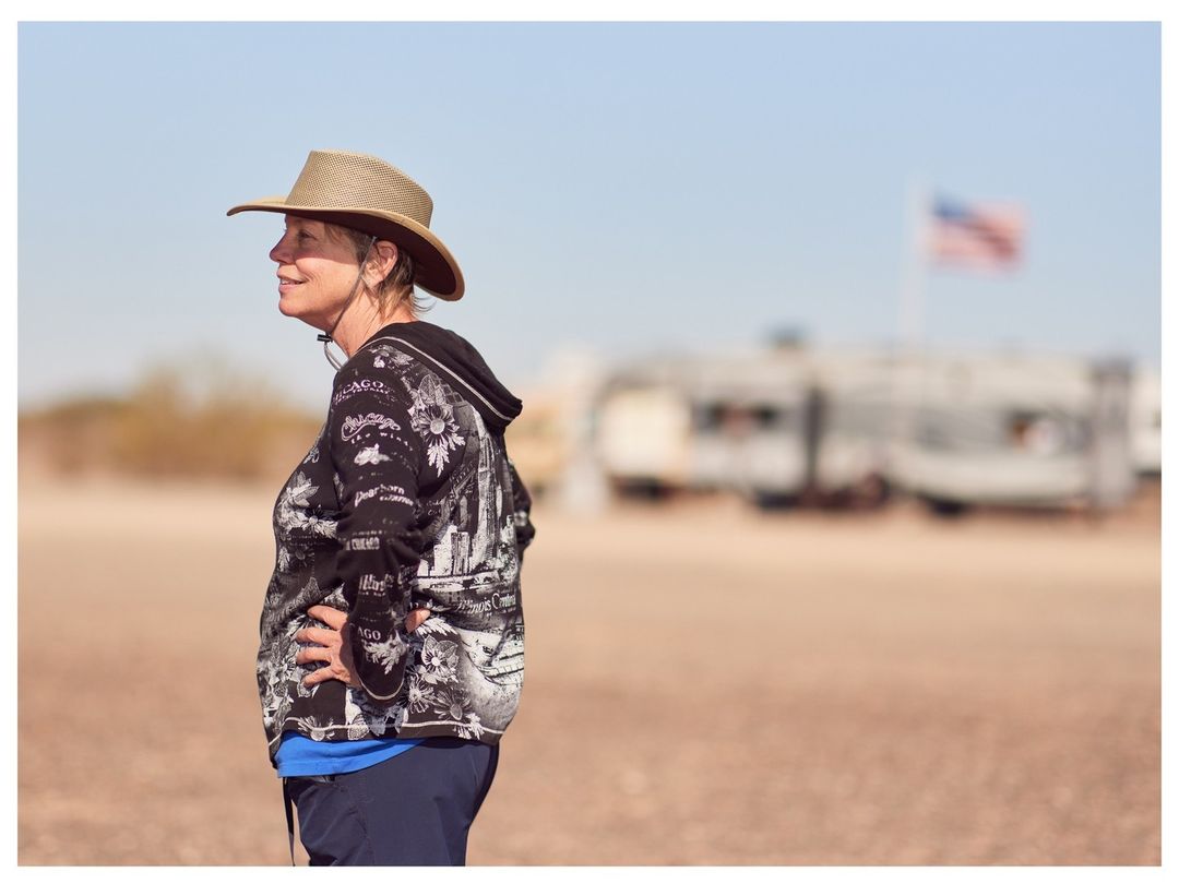 Adult female in a sunhat outside on a sunny day in an open space, wearing a hoodie, with recreational vehicles and an American flag in the background.