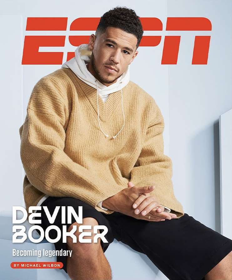 Devin Booker poses for a cover shot, wearing a tan sweater over a white hoodie, while seated against a white wall with an ESPN logo.
