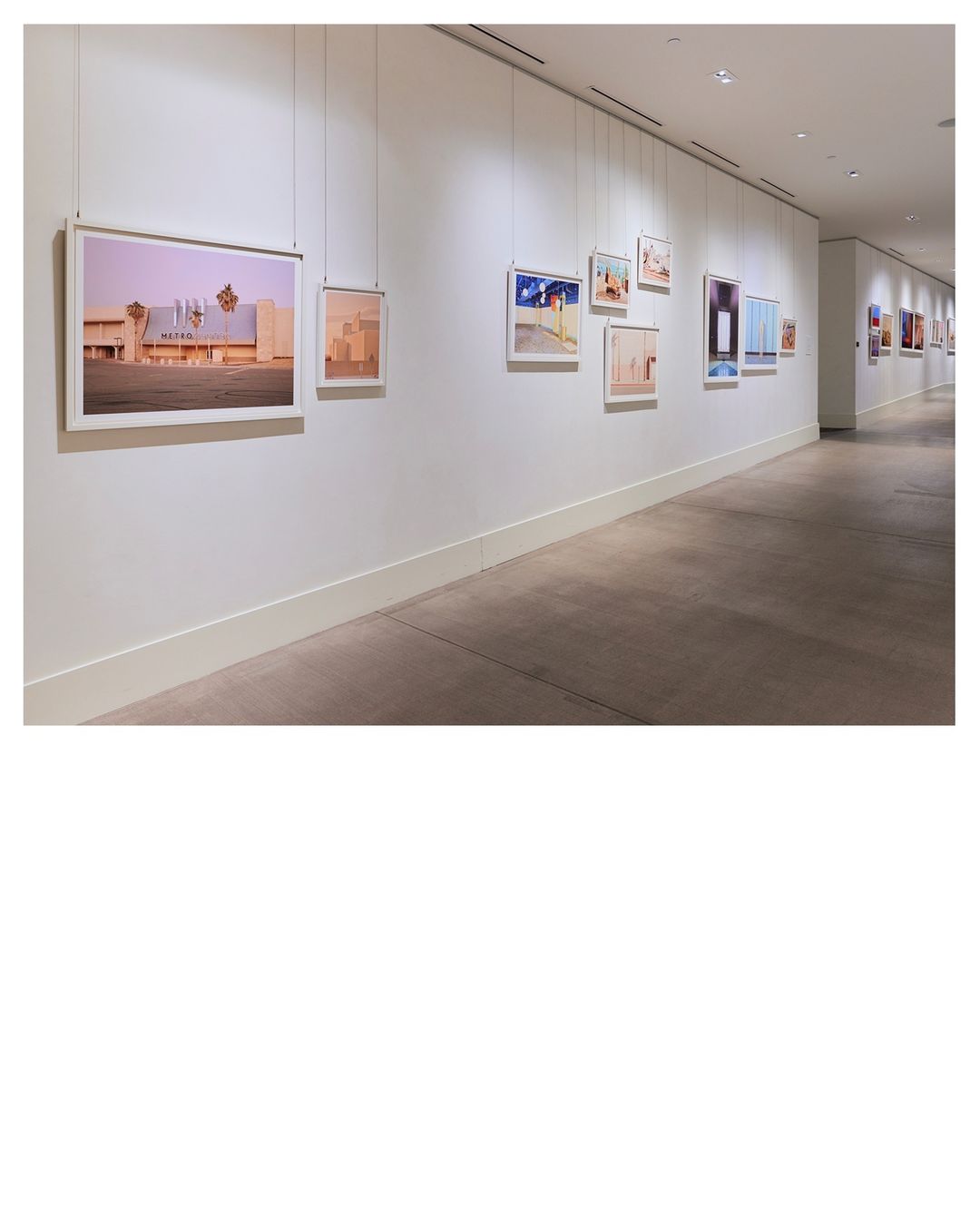 Art gallery featuring framed photographs hanging on a white wall along a corridor with gray flooring and bright, diffused lighting.