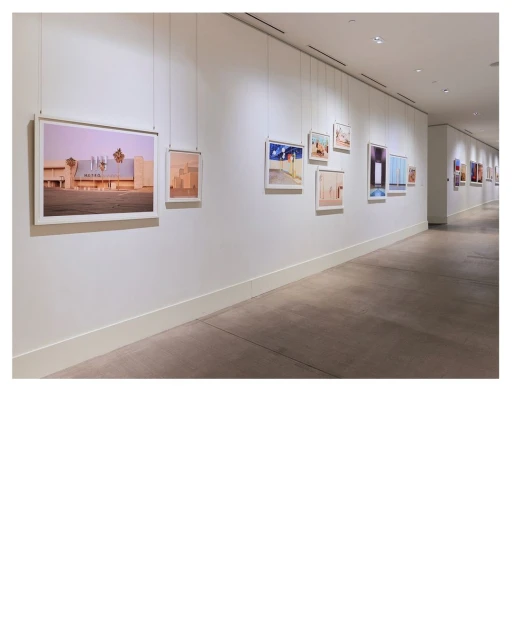 Art gallery featuring framed photographs hanging on a white wall along a corridor with gray flooring and bright, diffused lighting.