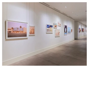 Art gallery featuring framed photographs hanging on a white wall along a corridor with gray flooring and bright, diffused lighting.