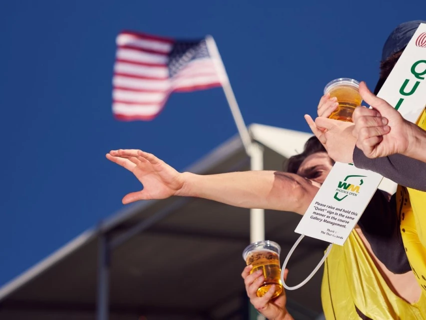 Spectators at a golf event hold beers and a 'Quiet' sign, with a blurred American flag waving against a clear blue sky backdrop.
