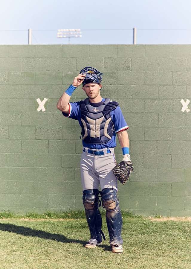 Baseball catcher in uniform, mask in hand, standing on a grassy field in front of a green wall on a sunny day. Full shot.