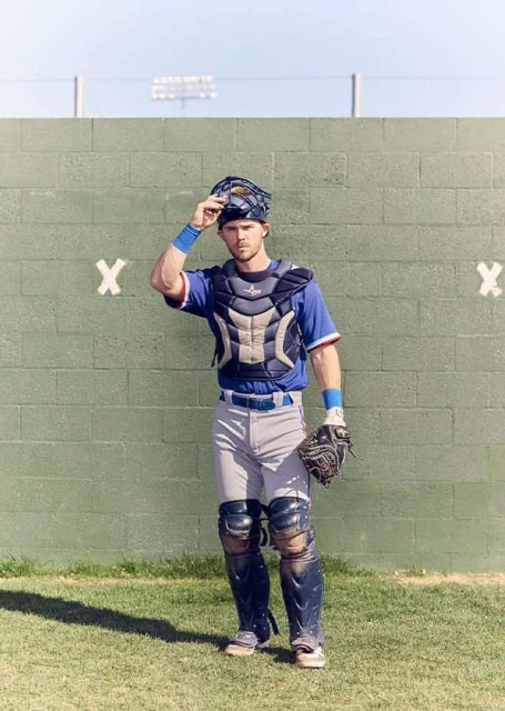 Baseball catcher in uniform, mask in hand, standing on a grassy field in front of a green wall on a sunny day. Full shot.
