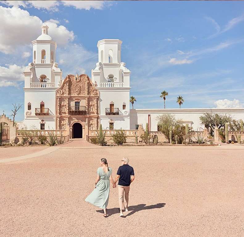 Couple strolling hand-in-hand towards the Mission San Xavier del Bac church with its striking white facade and ornate architectural details.