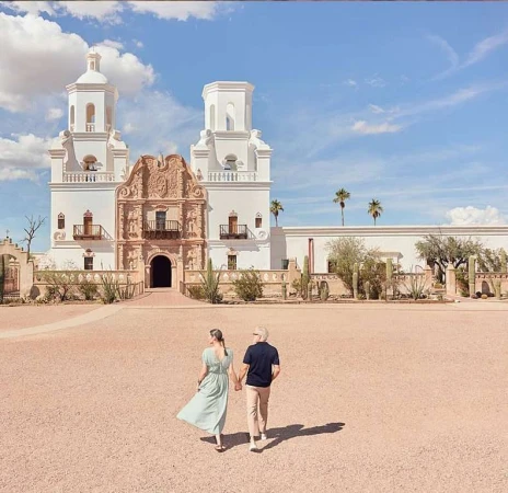 Couple strolling hand-in-hand towards the Mission San Xavier del Bac church with its striking white facade and ornate architectural details.
