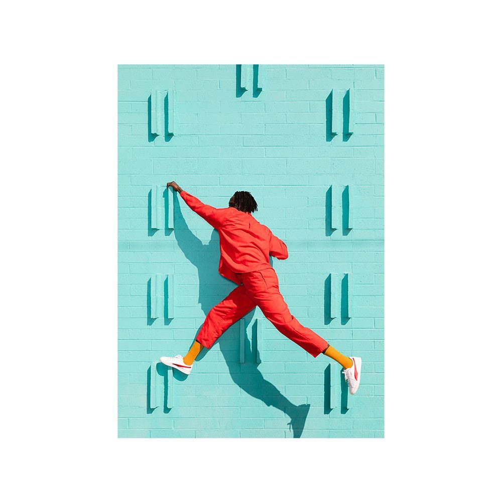 Athletic adult male leaps and reaches against patterned aqua wall wearing a red tracksuit, yellow socks, and white sneakers in bright sunlight.