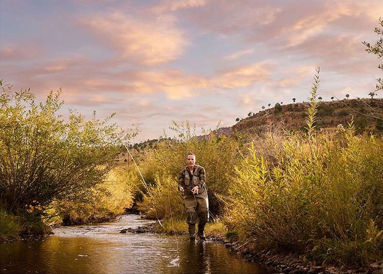 An adult male fly fishing in a shallow stream. The landscape includes tall reeds, bushes and a mountain range under a pastel sunset sky.