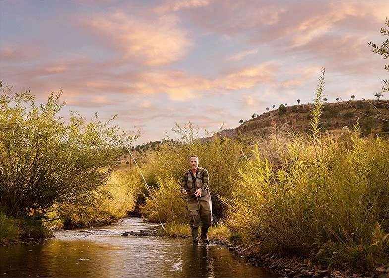 An adult male fly fishing in a shallow stream. The landscape includes tall reeds, bushes and a mountain range under a pastel sunset sky.