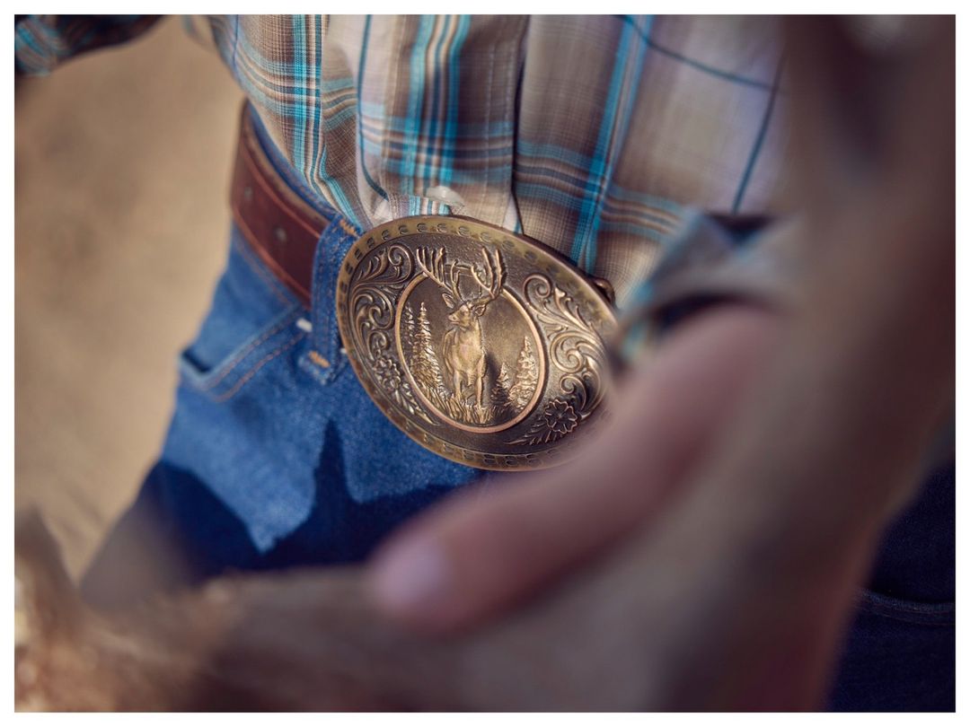 Person wearing a plaid shirt, blue jeans, and a large bronze belt buckle with a deer stands outdoors in a tan environment.