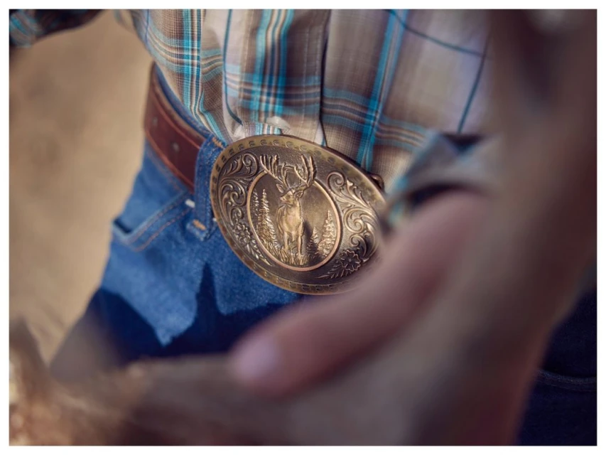 Person wearing a plaid shirt, blue jeans, and a large bronze belt buckle with a deer stands outdoors in a tan environment.