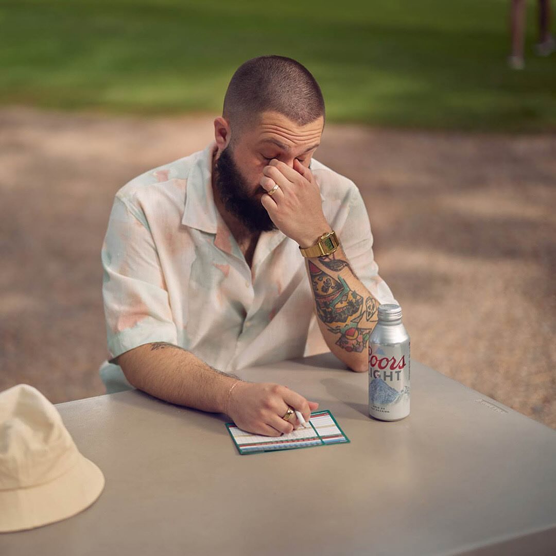 Adult male sitting at a table with a can of Coors Light, wearing a short-sleeve shirt, and a gold watch, resting his face in his hand.