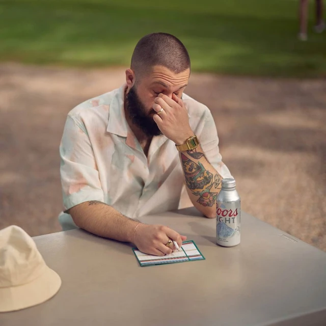 Adult male sitting at a table with a can of Coors Light, wearing a short-sleeve shirt, and a gold watch, resting his face in his hand.