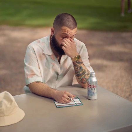 Adult male sitting at a table with a can of Coors Light, wearing a short-sleeve shirt, and a gold watch, resting his face in his hand.