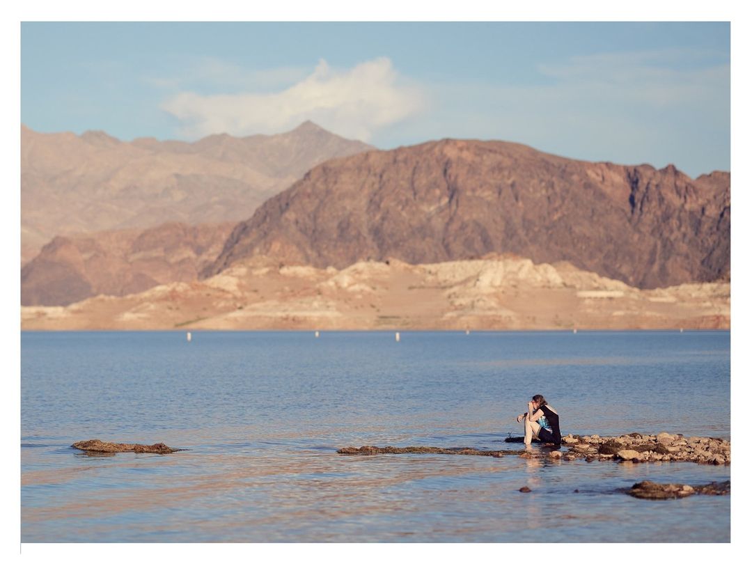 Couple sitting by a lake with mountains in the background. The water is calm and rocks are visible in the shallow area.
