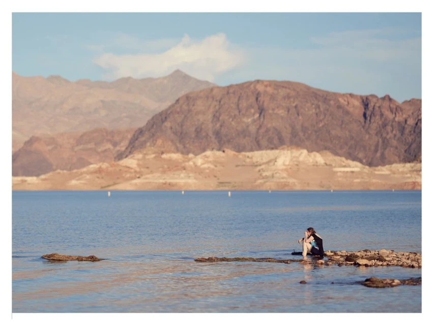 Couple sitting by a lake with mountains in the background. The water is calm and rocks are visible in the shallow area.
