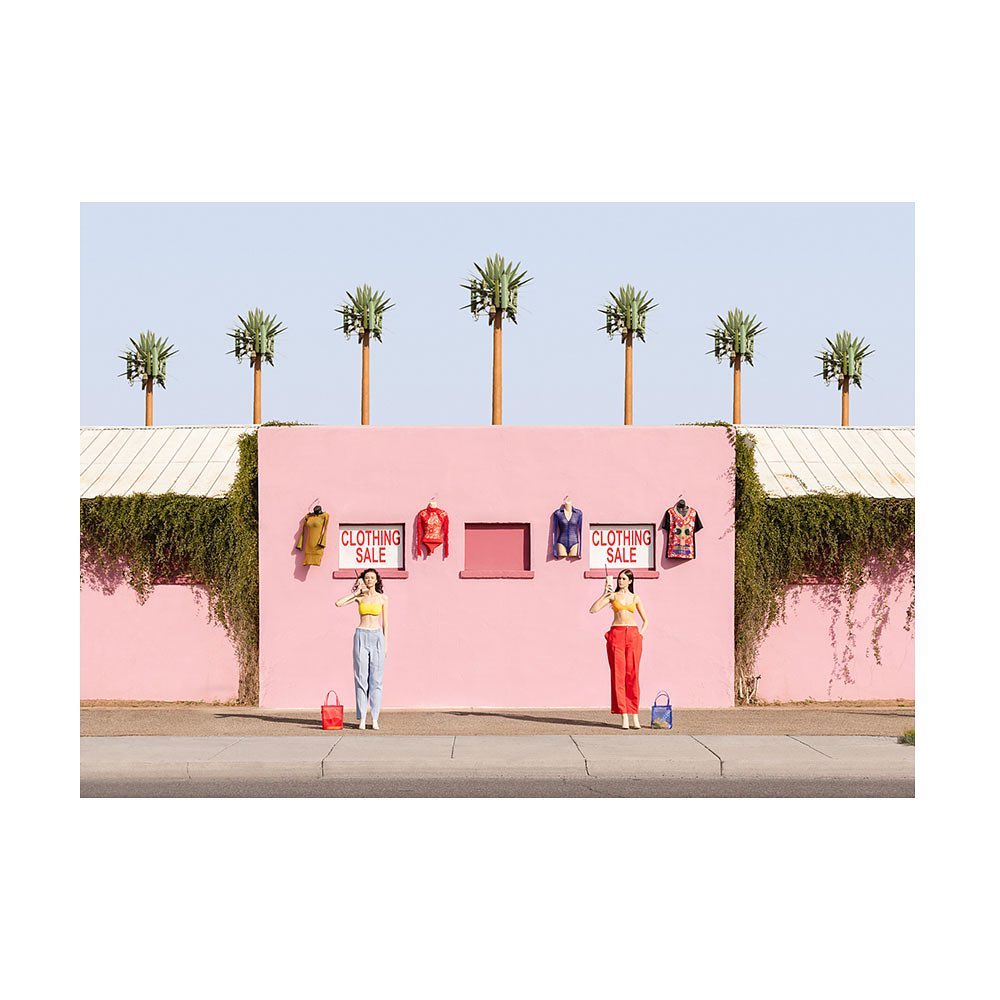Two women stand in front of a pink wall with clothes on display near palm tree cell towers, holding purses and wearing yellow tops.