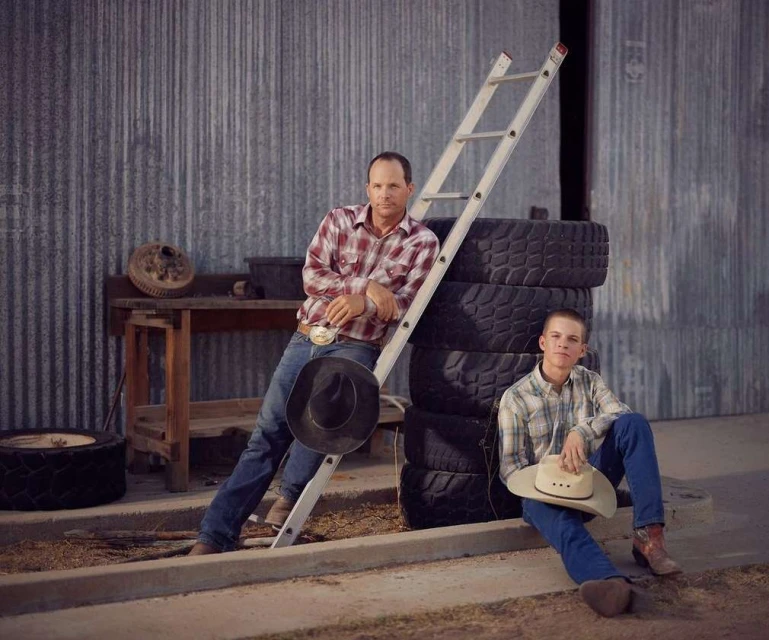 Two males in Western attire are sitting and leaning against a ladder and stacked tires in front of a metal corrugated wall with tools nearby.