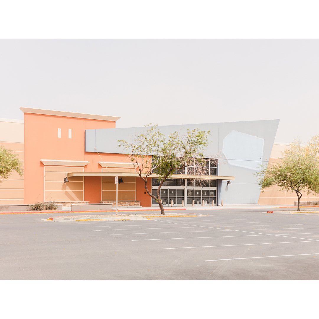 Deserted retail building on a sunny day, showcasing modern architecture, an empty parking lot, and sparse trees against a pale sky.