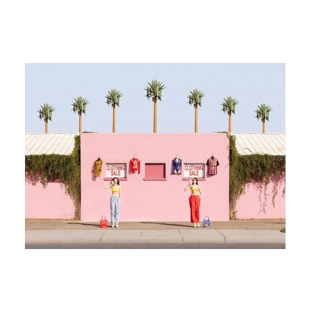 Two women posing in front of a bright pink building with clothing sale signs and palm trees under a clear blue sky
