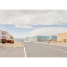 An empty road curving past pastel-coloured industrial buildings and desert shrubs under a pale overcast sky.