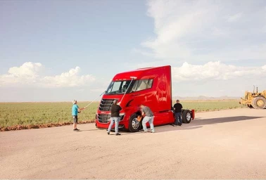 Four people cleaning and polishing a large bright red semi truck parked on an open dusty road under a clear sky.