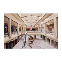 Interior shot of an empty abandoned shopping mall atrium with an escalator, vacant storefronts, and an American flag hanging from the ceiling.