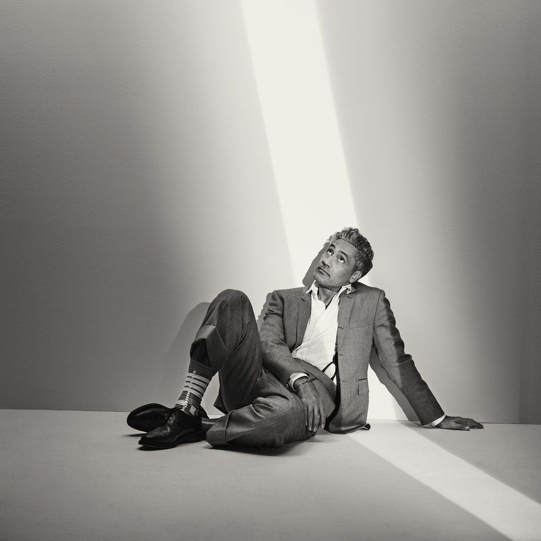 Monochromatic portrait of a man in a suit sitting and looking up toward a beam of light with patterned socks and black dress shoes.