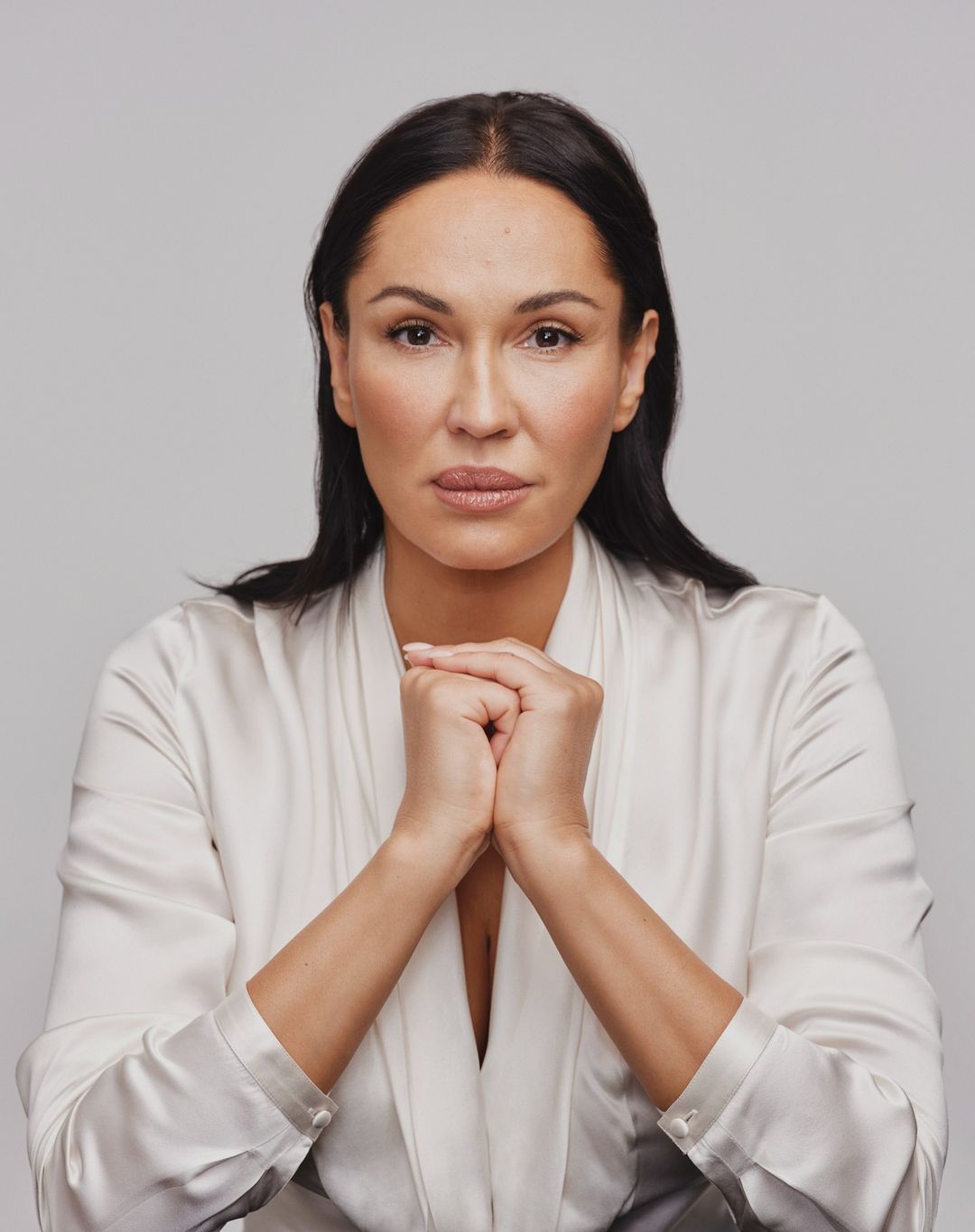 Close-up portrait of an adult woman with dark hair, wearing a light-colored satin robe, with her hands clasped together. Neutral background.