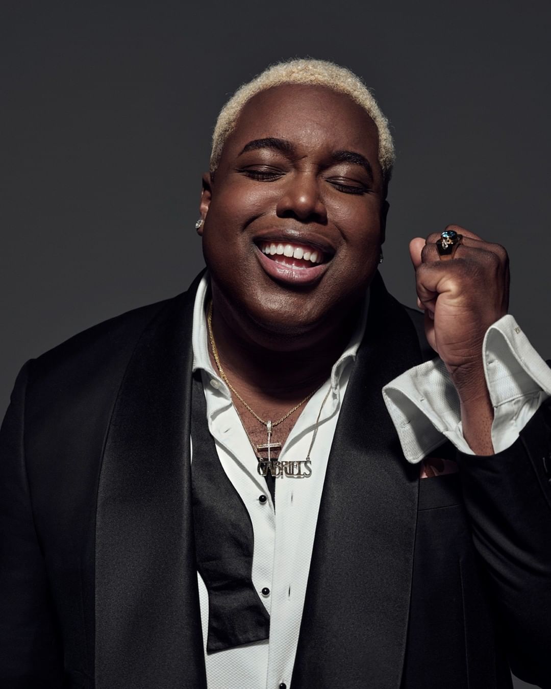 Close-up studio shot of an adult male with short blond hair, wearing a black tuxedo jacket, white shirt, jewelry, and smiling.