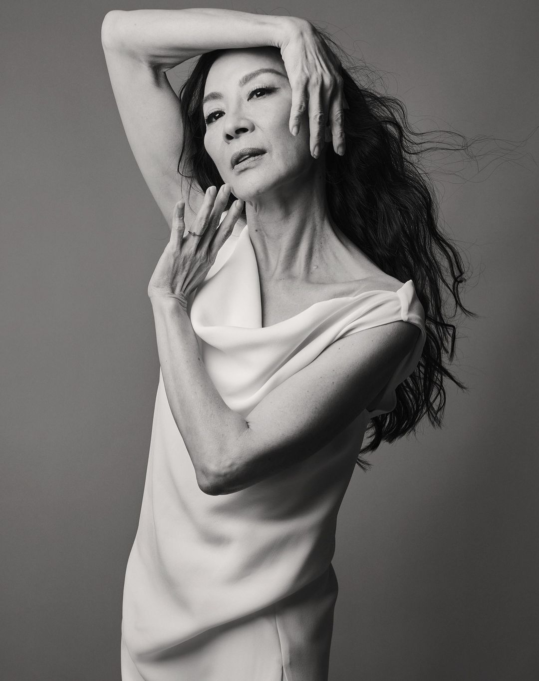 Elegant monochrome portrait of Michelle Yeoh in a draped dress, posed against a gray backdrop with soft, diffused lighting.