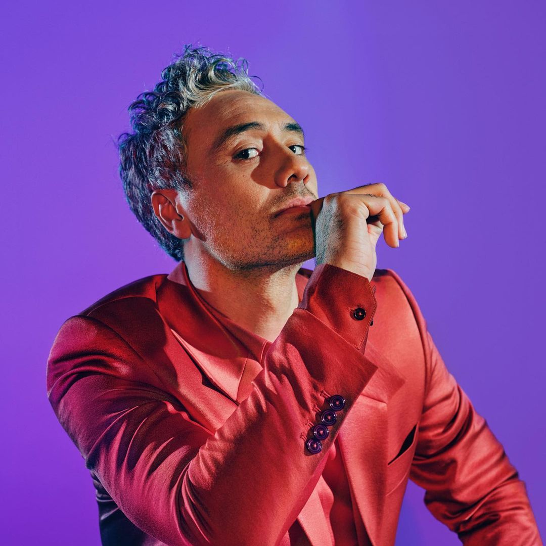 Close-up portrait of an adult male with graying curly hair, wearing a red blazer, posing against a purple studio backdrop with moody lighting.