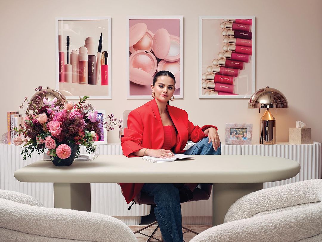 Selena Gomez in a red blazer sits at a desk with makeup posters behind her in a well-lit room with neutral colors and flower arrangement.