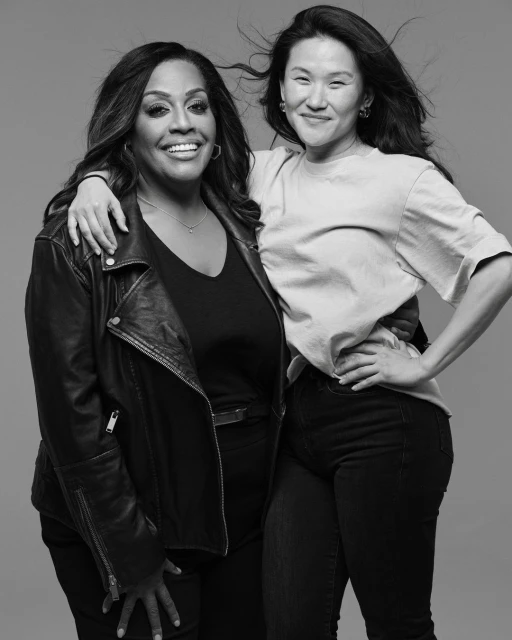 Black and white portrait of two adult women smiling while posing indoors. One woman has a leather jacket, the other a crew neck shirt.