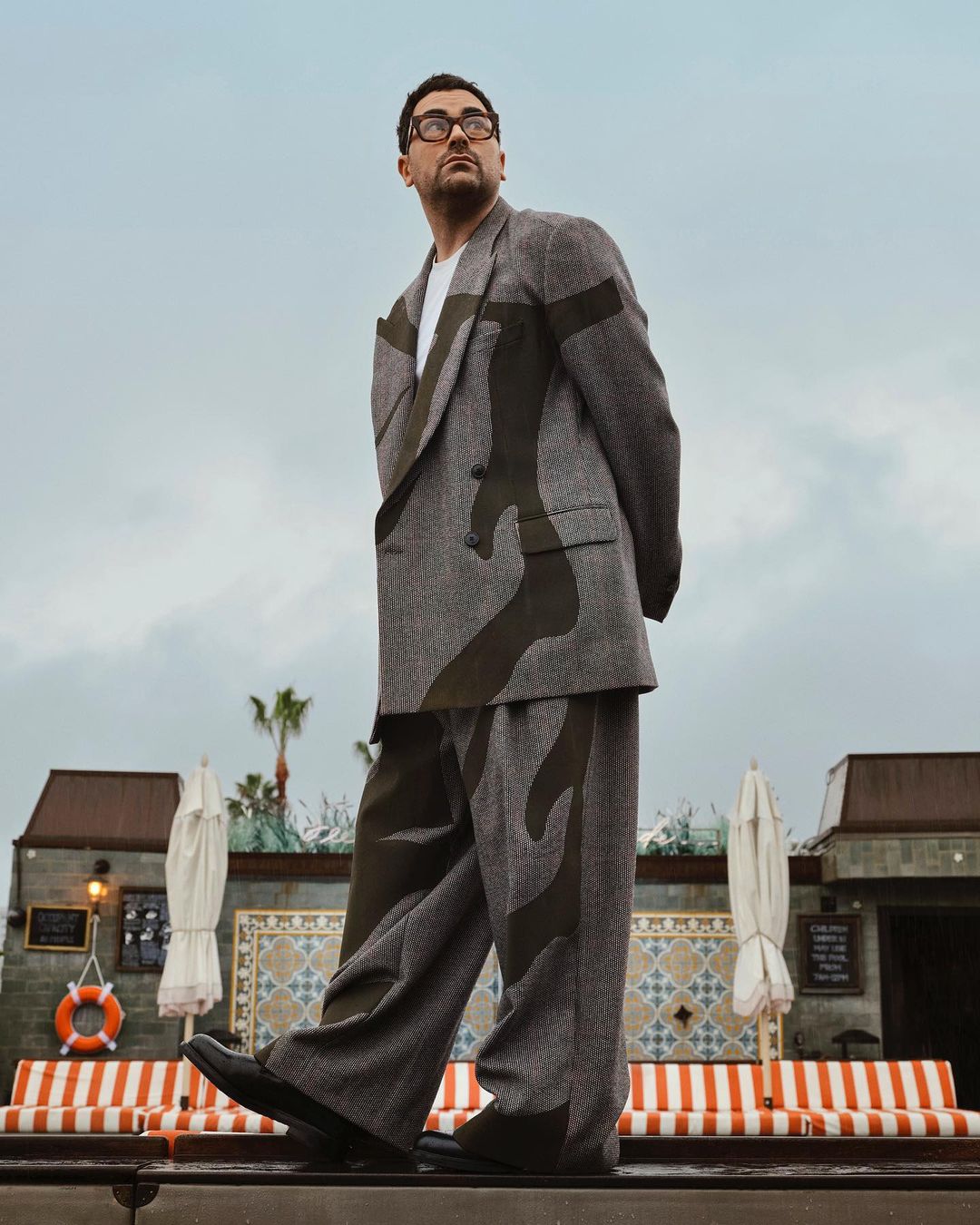 Man in oversized grey suit with graphic detail standing outdoors near building on sunny day. Cloudy sky, palm tree in the background.