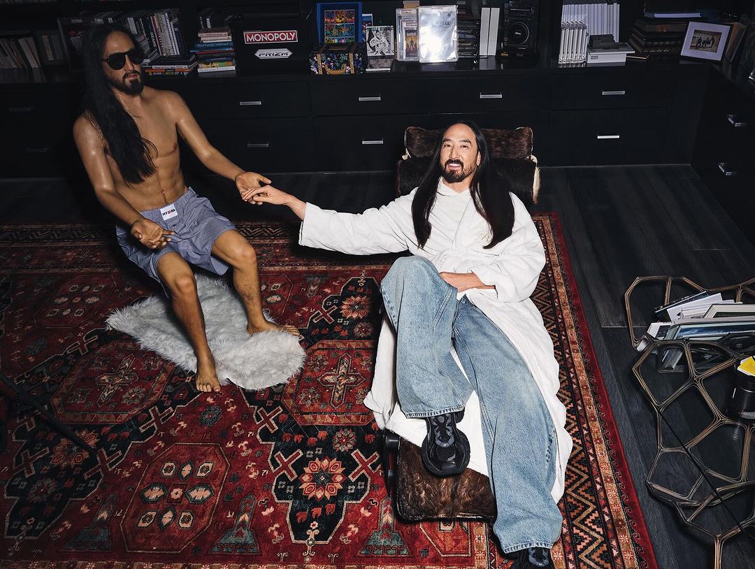 Steve Aoki sits next to his wax figure in a dark room. Aoki wears a white robe and jeans while the wax figure is shirtless.