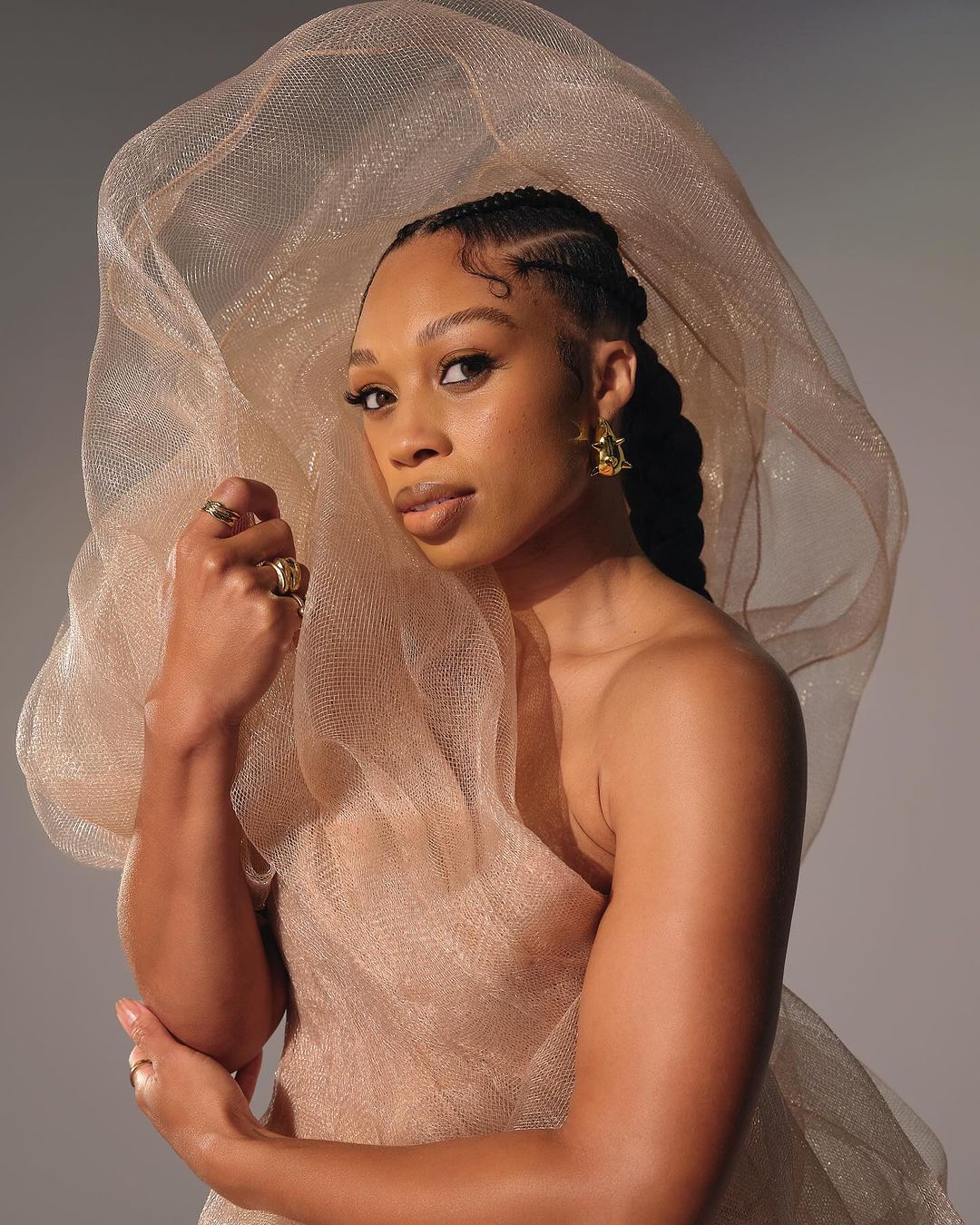 Portrait of a Black woman with elegant braided hair, posing with a neutral facial expression, wearing gold jewellery and a beige gown.