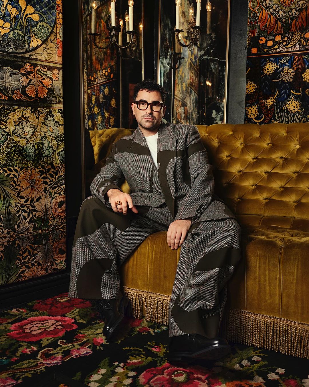 Adult male with glasses in gray patterned suit seated on a gold tufted couch in an ornately decorated room with floral patterned carpet.