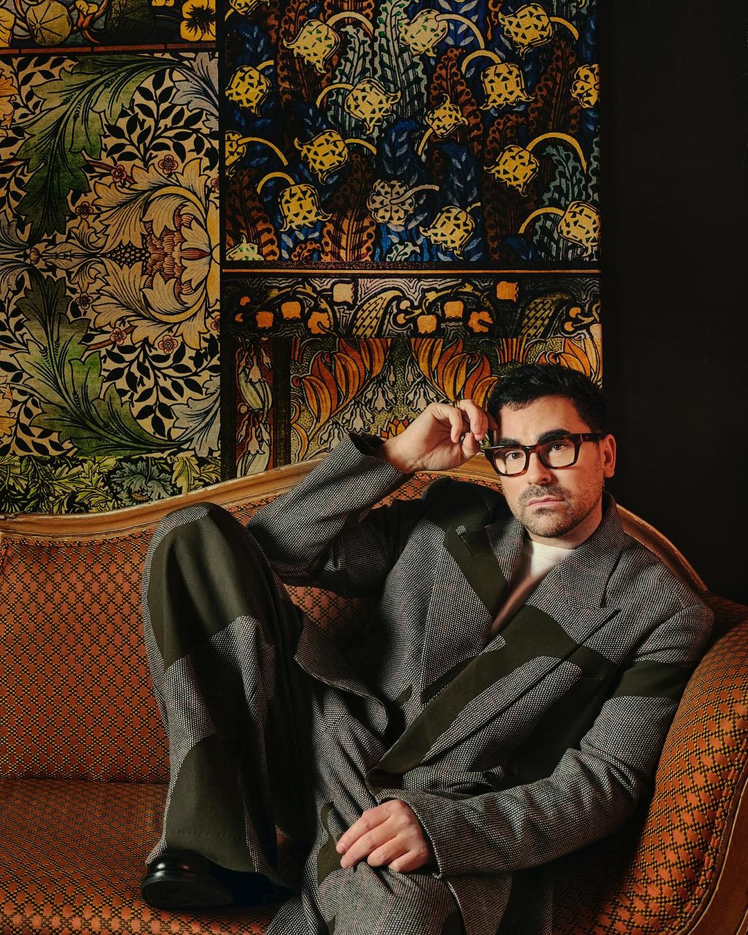 Portrait of a man with glasses in a patterned suit, reclining on an orange couch with patterned wallpaper in the background.