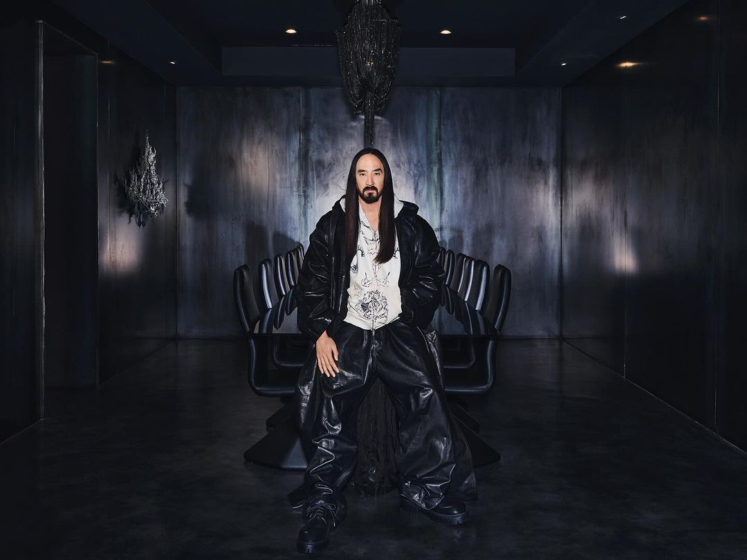 Steve Aoki in an indoor studio wearing leather pants, jacket and white hoodie posing in front of a metal wall and black chairs.