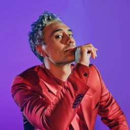 Portrait of Taika Waititi wearing a red satin blazer against a solid purple background