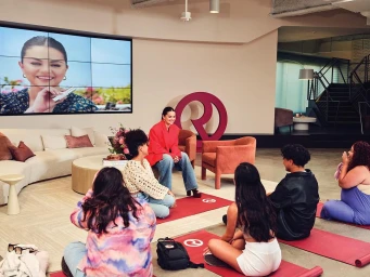 Selena Gomez in a red blazer sits on a table addressing a group of people seated on yoga mats in a modern studio space by Jessica Chou.