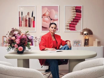 Selena Gomez wearing a red blazer seated at a pale curved desk with three framed beauty product prints on the wall behind her