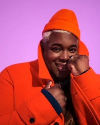 Portrait of a smiling Black man wearing a bright orange beanie and matching wool coat against a solid pink background.