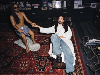 Man in white robe and blue jeans sitting on chaise lounge holding hands with a wax figure of himself in a dark room