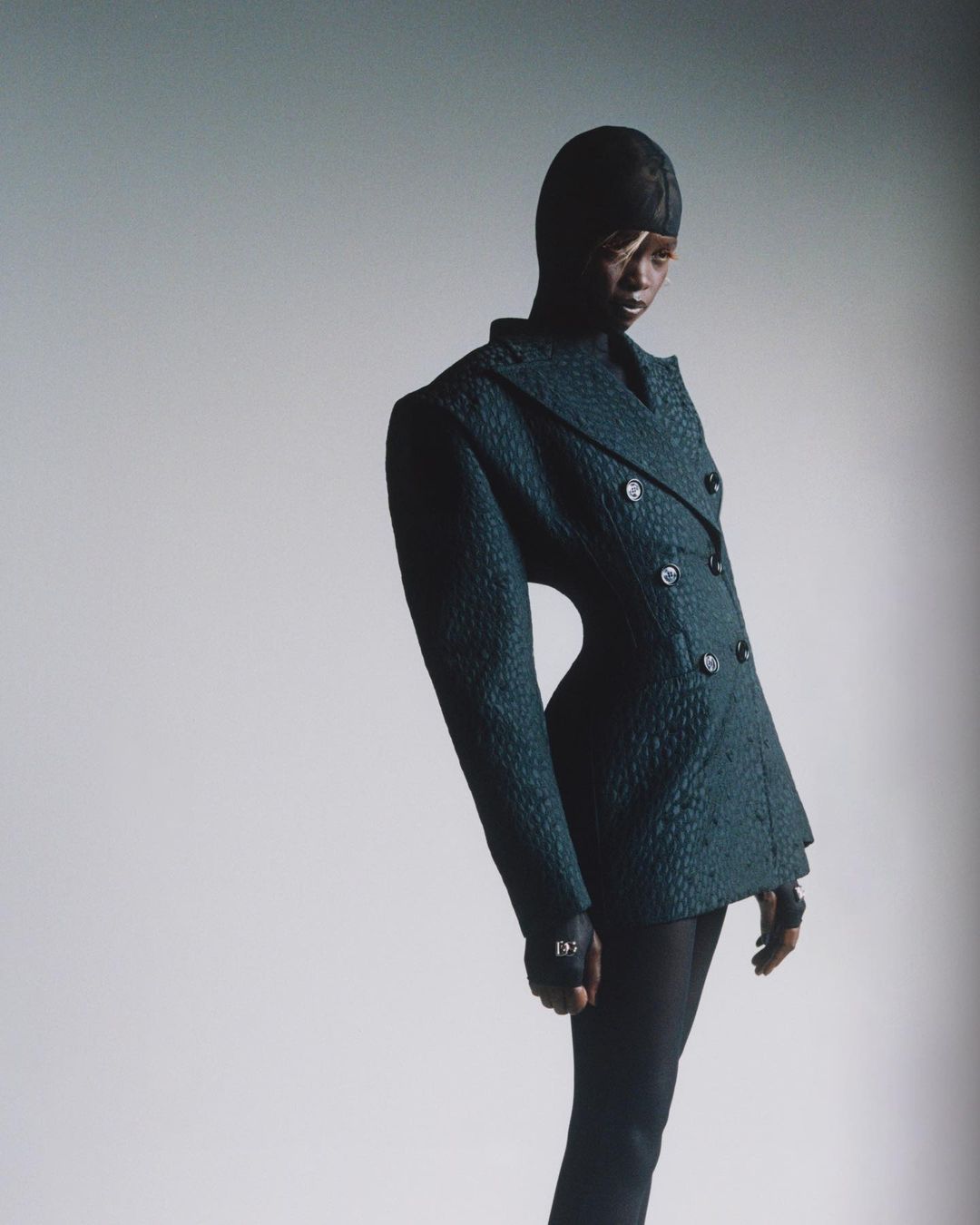 Model in textured dark green double-breasted blazer and head covering against a plain white studio wall