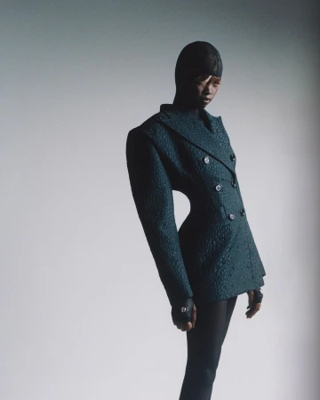 Model in textured dark green double-breasted blazer and head covering against a plain white studio wall
