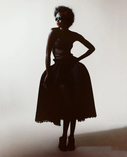A striking full-length studio portrait of a dark-skinned female fashion model wearing a vintage black couture dress with a voluminous skirt.