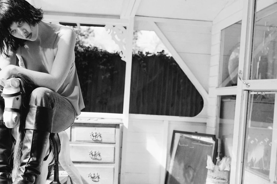 Monochrome portrait of a woman with dark hair seated on a toy horse inside a white wooden structure with an open window.